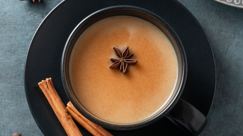 A black mug and saucer filled with chai and a star anise pod alongside whole cinnamon sticks