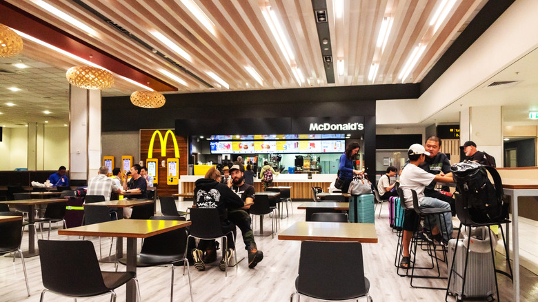 The interior of a McDonald's resturant dining area, facing the order counter. Several groups sit on chairs around tables or high top tables.