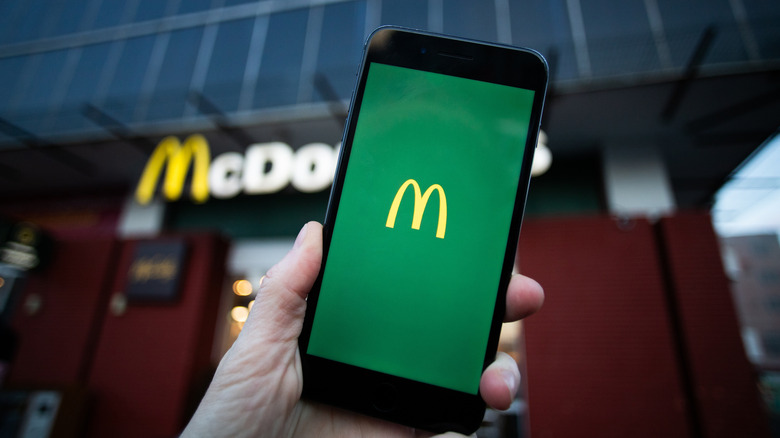 Hand holds smartphone in front of McDonald's storefront in background. A screen with the golden arches is open on the smartphone.
