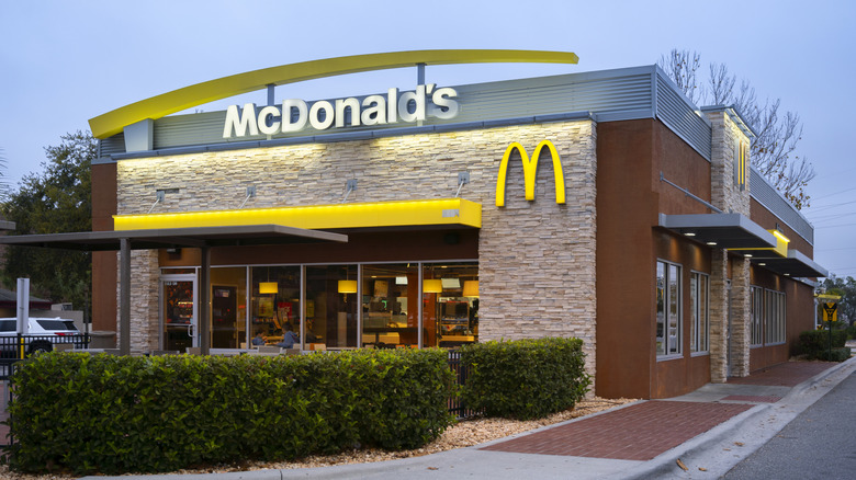 A McDonald's storefront with the golden arches and store logo on the front on stone facade. The picture is taken from the parking lot in front of the building.