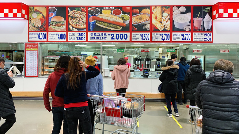 Customers line up at the food court inside a South Korean Costco, with menu items like bulgogi bakes, Kirkland hot dog set, chicken burgers, and mushroom soup displayed above the counter.