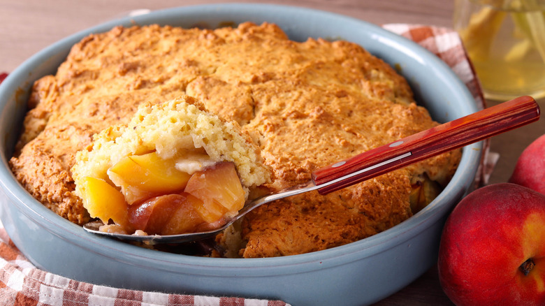 Homemade warm peach cobbler in a tin with a spoon and peaches on the side