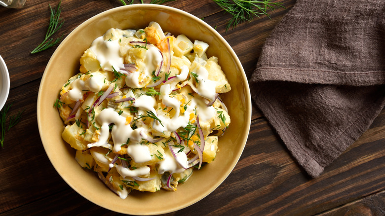 Top view of potato salad with egg, pickle, red onion with mayonnaise and yogurt dressing in bowl on wooden table.