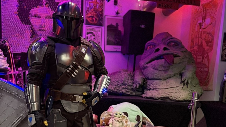 A person in a bounty hunter costume standing in front of a large Jabba the Hutt statue and a Grogu doll in 7 Stars Bar and Grill