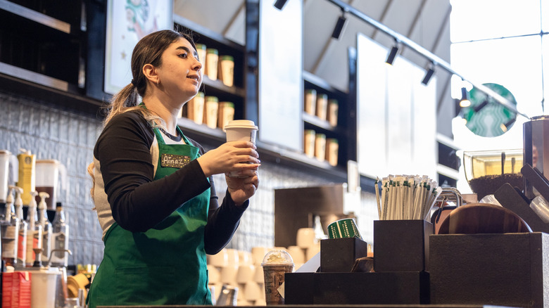 A Starbucks barista in a green apron holding a cup of coffee behind the counter.