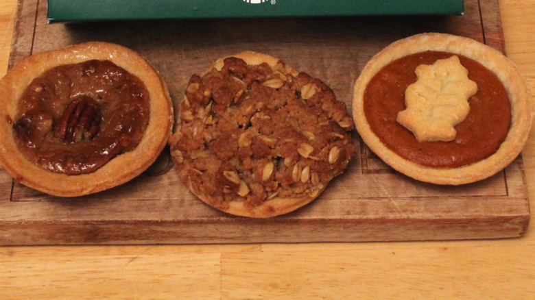 Starbucks' new Pecan, Apple Crumble, and Pumpkin mini pies on a wooden board in front of a branded box