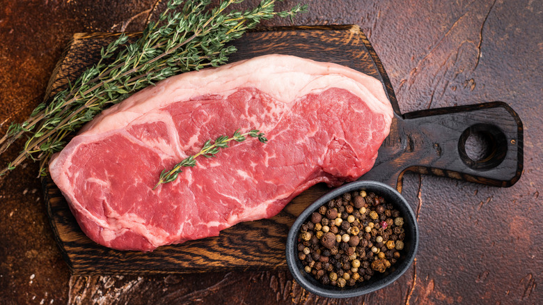 A raw strip steak on a wooden cutting board beside thyme and peppercorns.