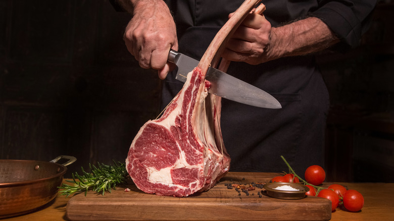 Man's hands cutting a steak with long bones that's standing on its end, on a wooden board with cherry tomatoes and rosemary springs against a dark background