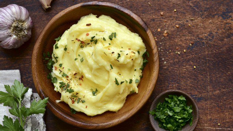 Mashed potatoes in a wooden bowl with fresh herbs and a bulb of garlic