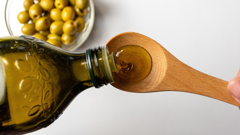 Pouring olive oil into a wooden spoon over a bowl of green olives