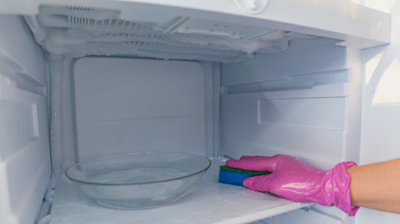 A person wearing a pink latex glove uses a sponge to wipe down a freezer interior