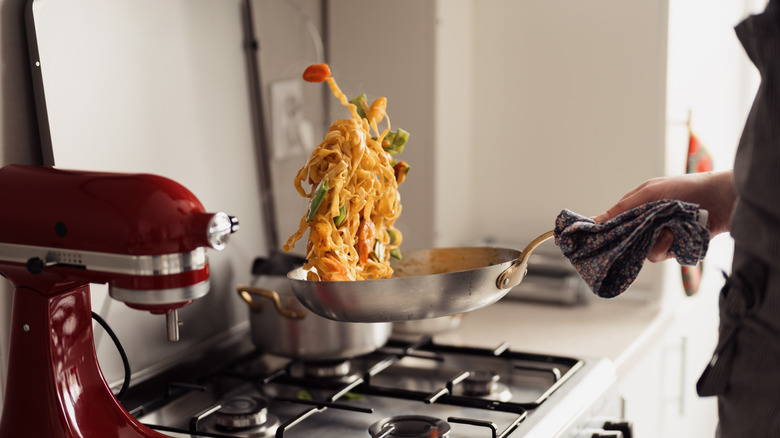 Someone tossing pasta over a stovetop in a metal frying pan