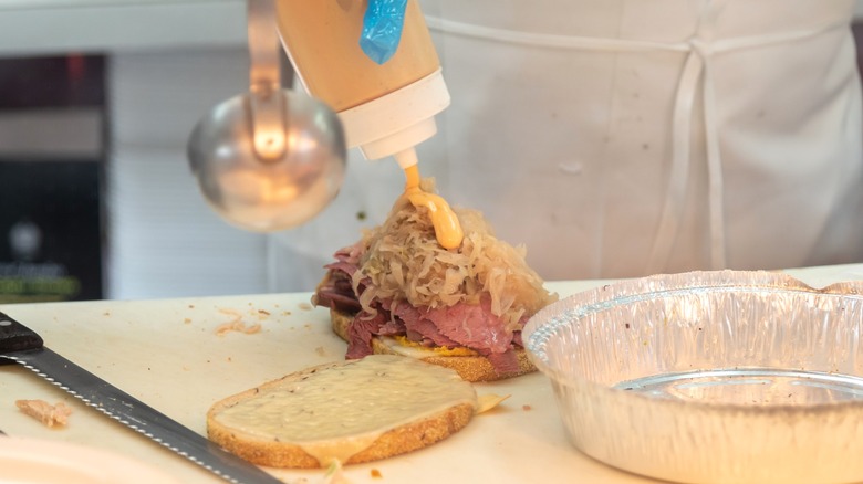 A chef squirts dressing onto sauerkraut in a Reuben sandwich
