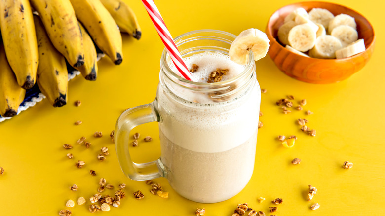 Banana smoothie in a glass jar mug with a red and white straw garnished with a banana slice on a yellow surface with scattered nuts with a small bowl of banana slices and other bananas in the background