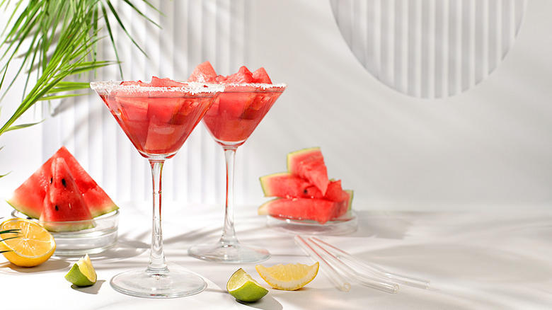 Tall martini glasses with frozen watermelon drink inside on a white table with citrus and watermelon slices nearby.