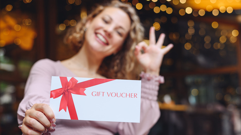 A smiling woman holding up a gift voucher envelope while giving an "okay" sign with the other hand