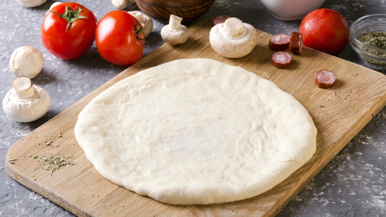 Raw pizza dough on a wood board with mushrooms and tomatoes