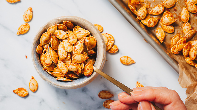 Overhead view of hand lifting a spoonful of pumpkin seeds, next to baking sheet