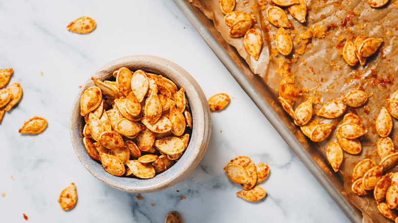 Overhead view of small bowl of pumpkin seeds, next to baking sheet