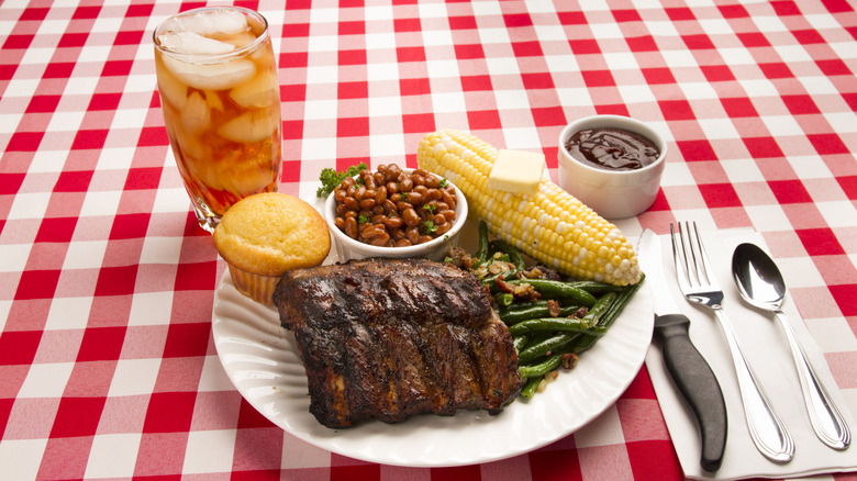 Plate of ribs, corn on the cob, beans, green beans, and cornbread with a glass of iced tea on a red checkered tablecloth