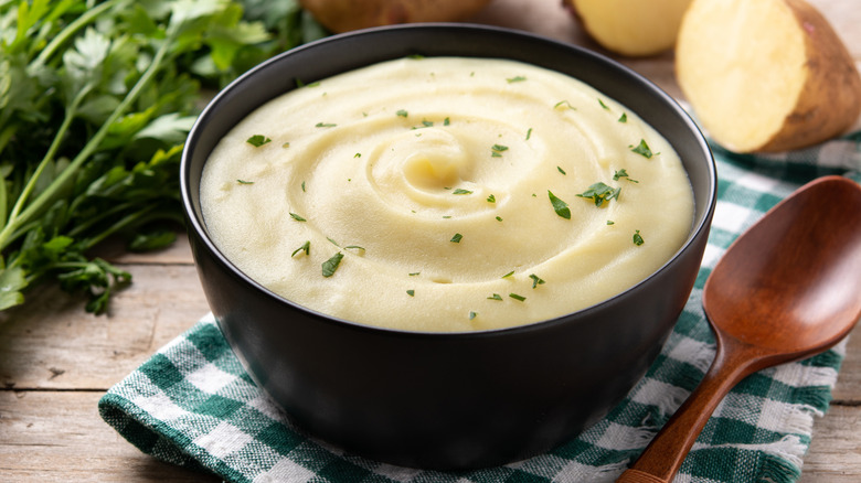 Mashed potatoes sprinkled with parsley bits in a small black bowl with a wooden spoon next to it on a green and white checkered cloth