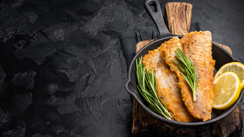 Fried catfish in a cast iron skillet with rosemary and lemon on a black background