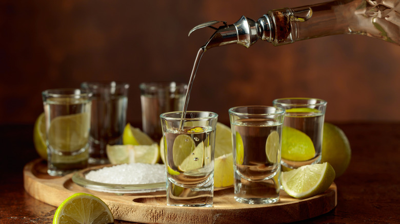 Gold tequila being poured into glasses with salt and lime slices on the side.