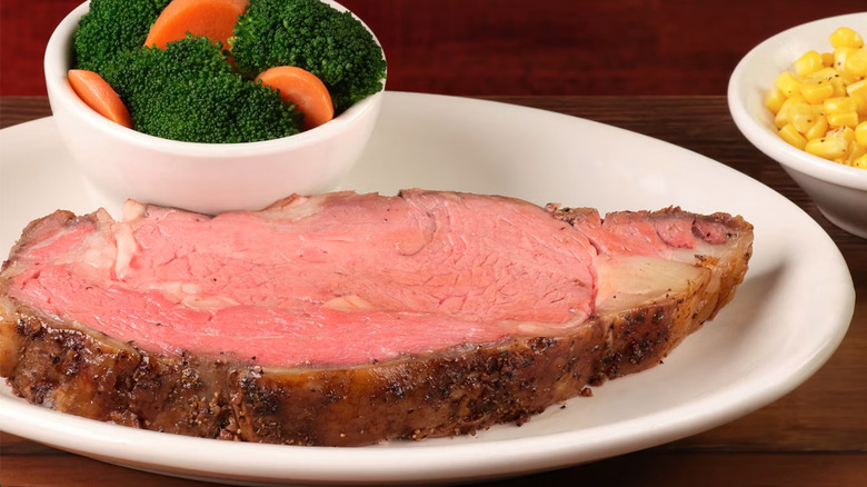Prime rib on white plate alongside bowl of steamed broccoli and carrots and bowl of corn