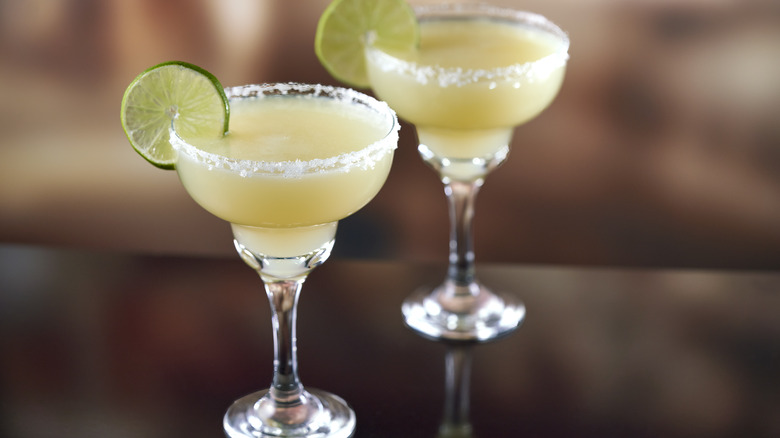 Two standard margaritas in margarita glasses with limes and sugar on the rim.
