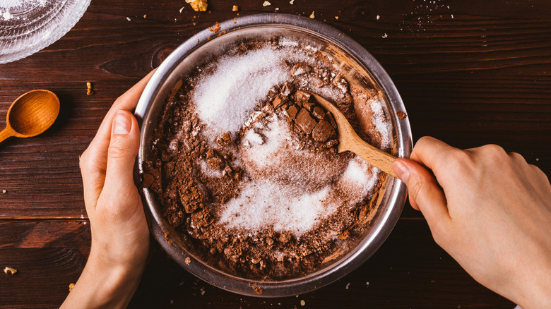 Mixing cocoa powder and sugar in a metal bowl, with a spoon, on a wooden surface