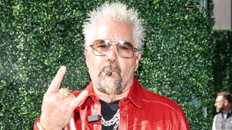 Guy Fieri in red leather jacket, smoking a cigar in front of greenery