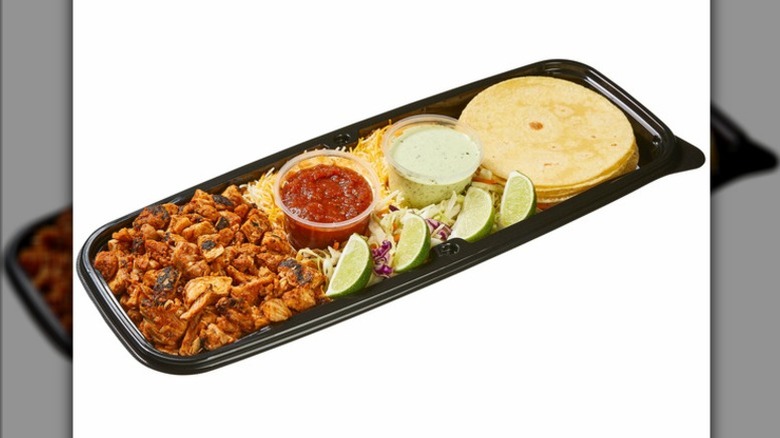 The chicken street taco from Costco, with corn tortillas, lime wedges, shredded cheese, seasoned ground meat, salsa, and crema on platic tray