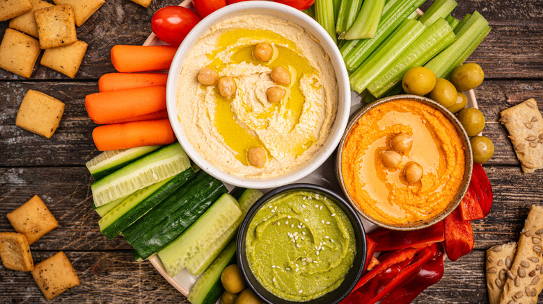 3 varieties of hummus in bowls on a wooden platter surrounded by sliced carrots, celery, cucumber, and crackers