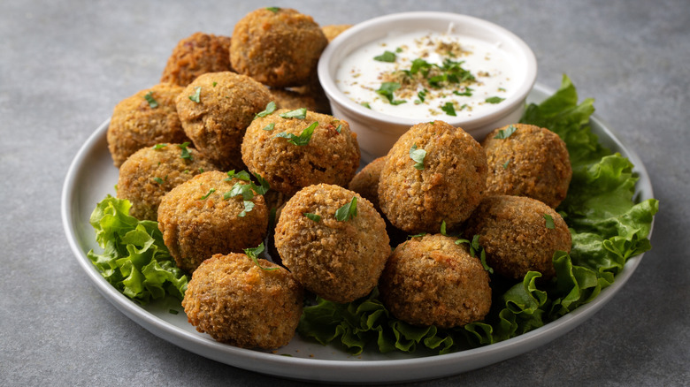 Crispy balls of falafel arranged on a plate over a bed of lettuce and with a bowl of creamy dipping sauce