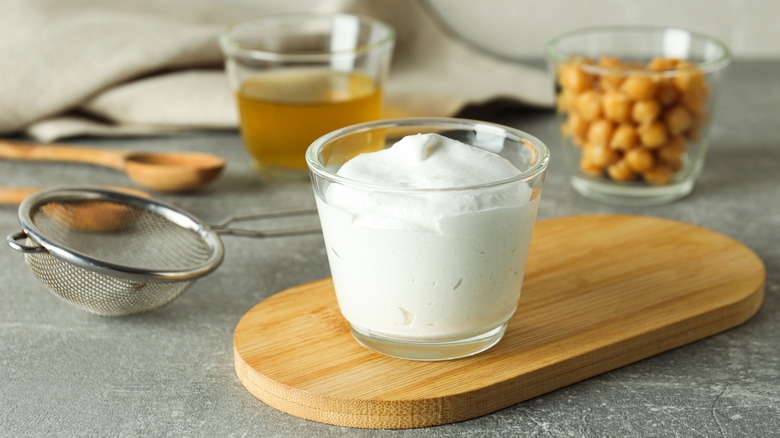 A glass bowl of frothy white cream on a wooden cutting board with a bowl of chickpeas in the background