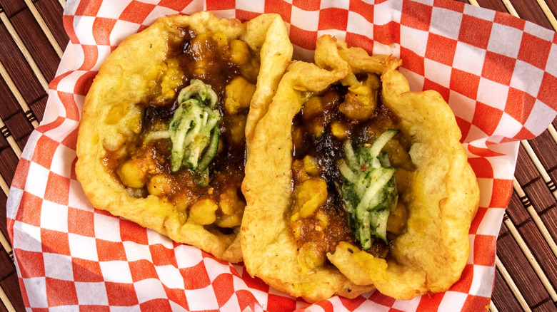 Two fried pieces of flatbread topped with curry chickpeas in basket with red and white checkered paper