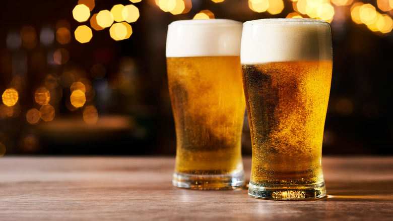 Two glasses of beers sitting on a beer with lights in the background.