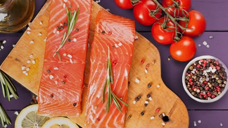 Two seasoned Kroger salmon fillets on a wooden cutting board near a bowl of peppercorns, cherry tomatoes, and lemon slices.