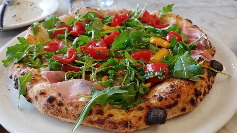 A whole Neapolitan-style pizza with prosciutto, arugula, and red and yellow tomatoes on a white place set on a white marble surface