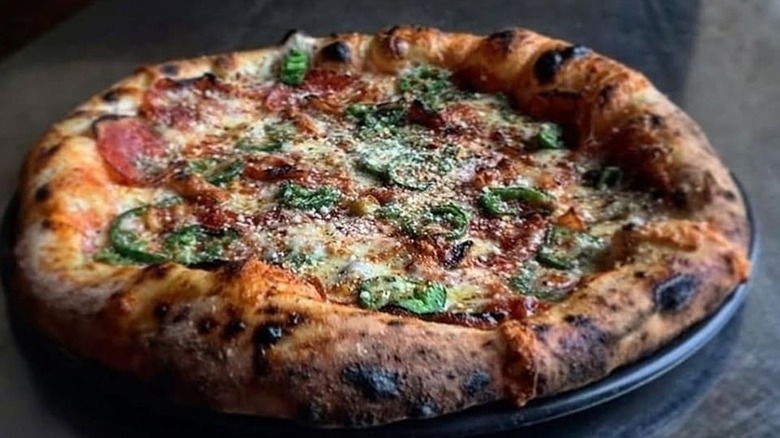 A whole Neapolitan-style pizza topped with soppressata, green peppers, mozzarella cheese, and parmesan on a black plate set on a gray surface