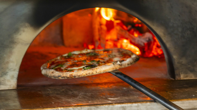 A Neapolitan-style pizza is removed from a hearth on a pizza peel with wood burning in the background