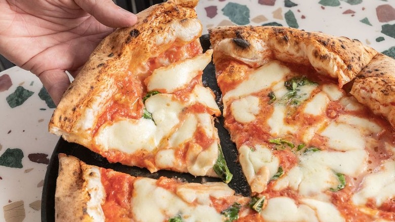 A hand (left), removing a slice from a whole Neapolitan-style Margherita pizza on a black plate set on a terrazzo surface
