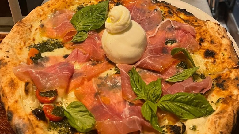 Close-up of a whole Neapolitan-style pizza topped with pesto, cherry tomatoes, prosciutto, and fresh mozzarella with a dollop of burrata in the middle