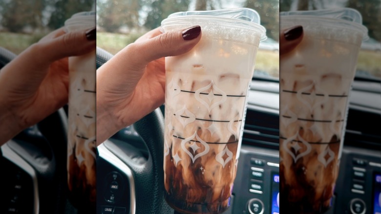 A hand holds a Starbucks espresso macchiato in a car