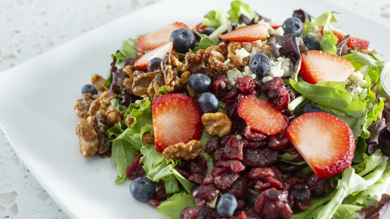Strawberries, blueberries, cranberries, and walnuts on a salad on a square white plate