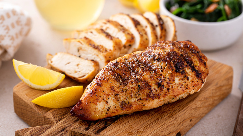 One whole and one sliced cooked chicken breast on a cutting board with sliced lemons