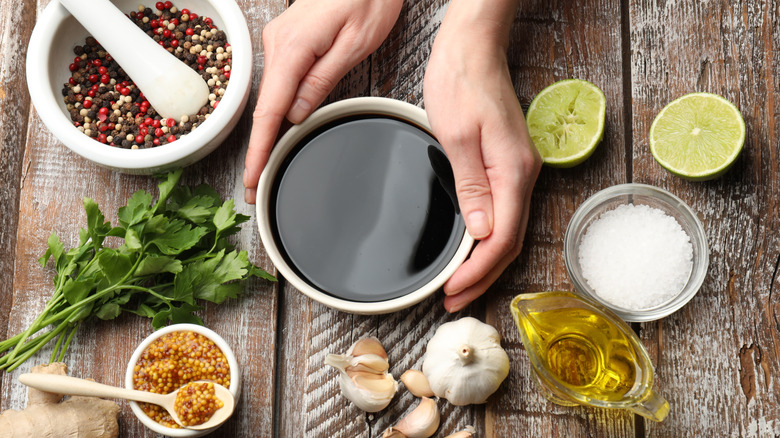 Various marinade ingredients on a wood table including soy sauce, salt, peppercorns, mustard seed, garlic, oil, lime halves, and parsley