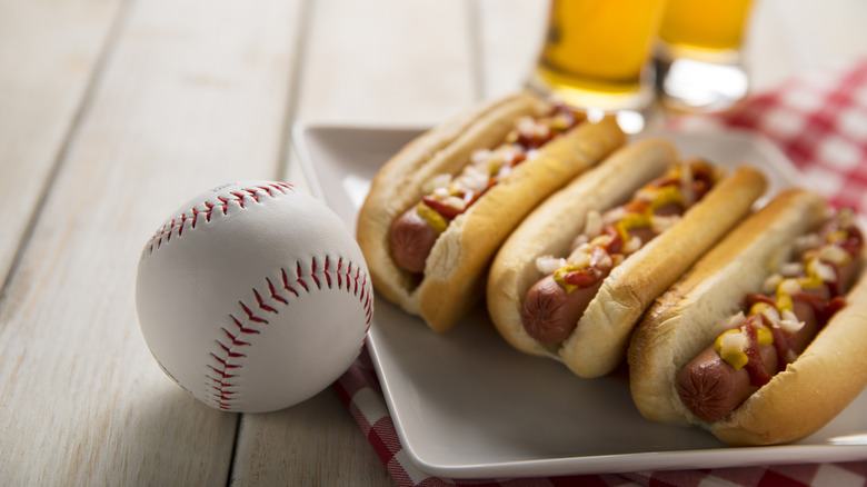 3 hot dogs topped with condiments on a tray, next to a baseball, with pints of beer in the background