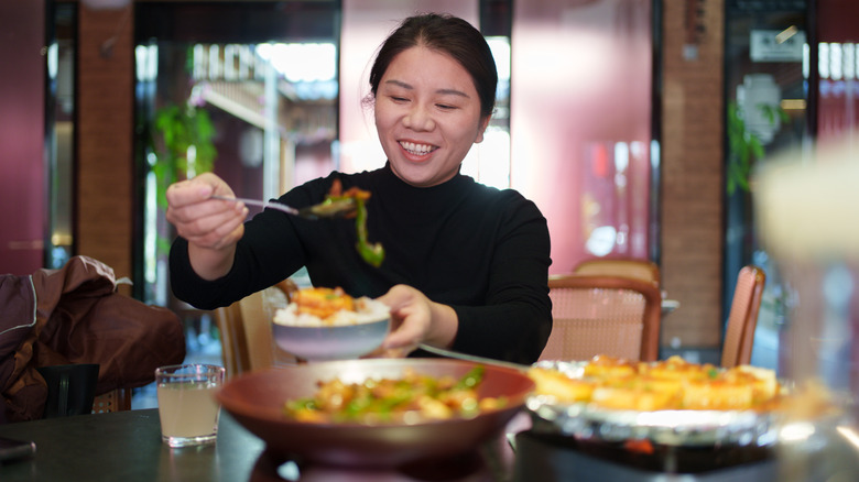 A smiling woman in a black long-sleeved shirt helping herself to more Chinese food.