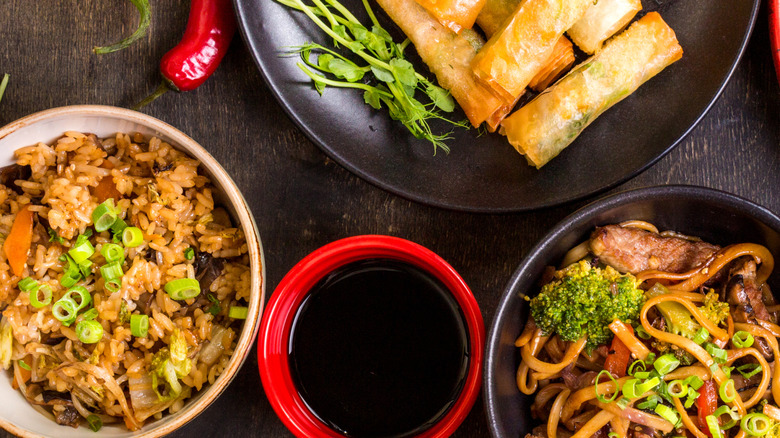 Multiple bowls and plates full of Chinese food placed on a wooden surface. There's rice, noodles, dumplings, and more.